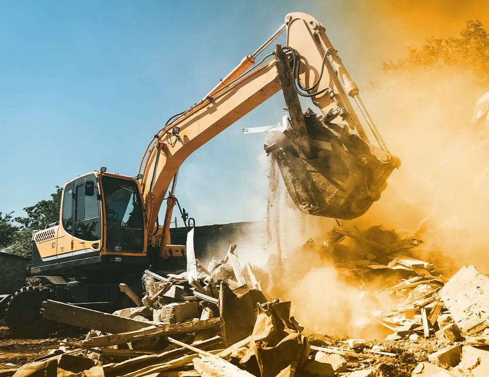 An excavator undertakes a small demo, using its bucket to lift debris from a demolished building amidst a cloud of dust under a clear blue sky.