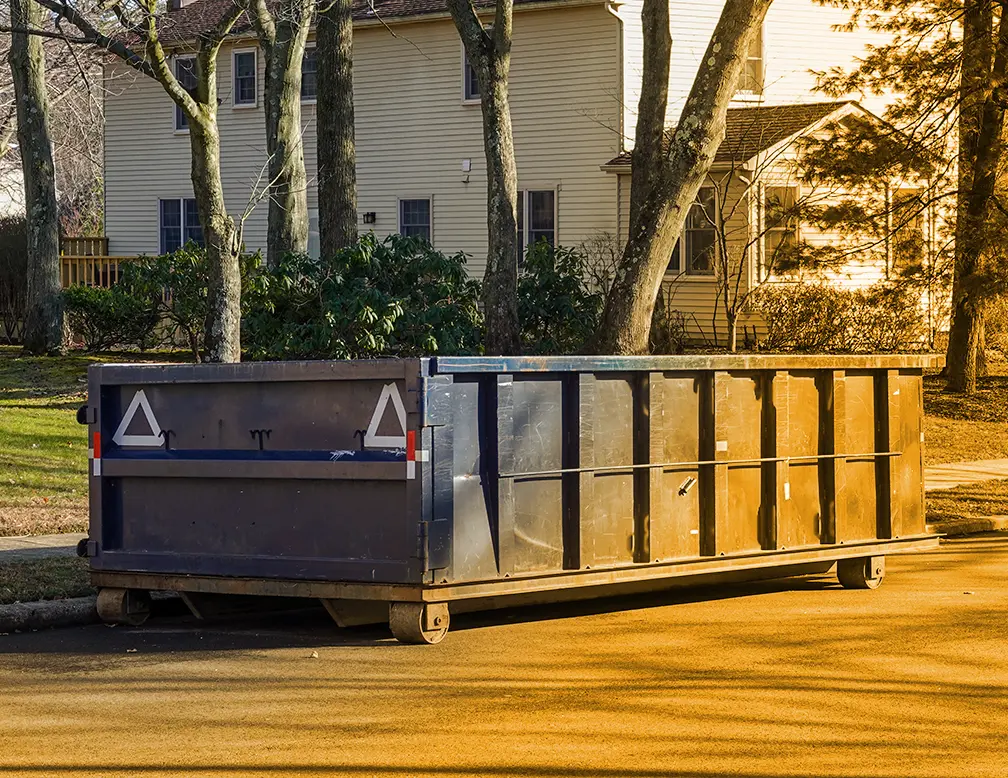 A large metal dumpster, likely earmarked for a pool removal or demolition project, is placed on the side of a residential street in front of a beige house with trees and shrubbery in the background.