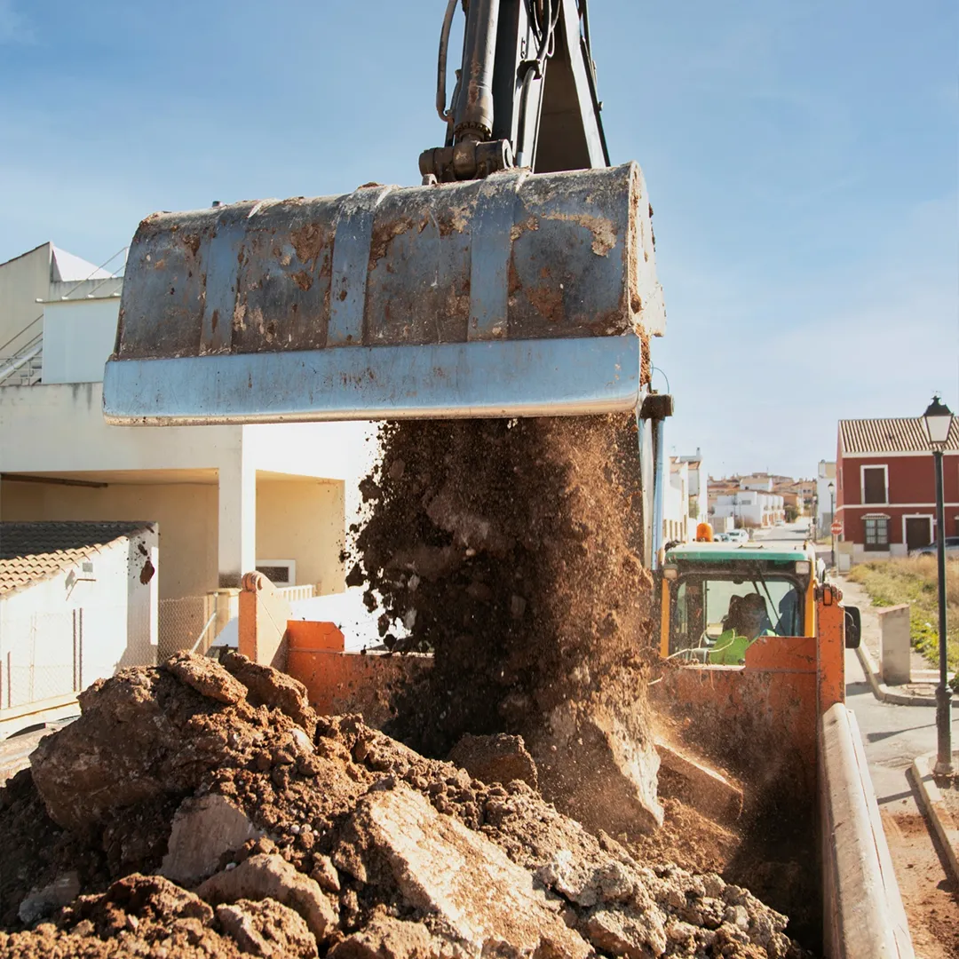 An excavator is busy with a pool removal, dumping a load of dirt and rocks into a truck on a construction site in a residential area.