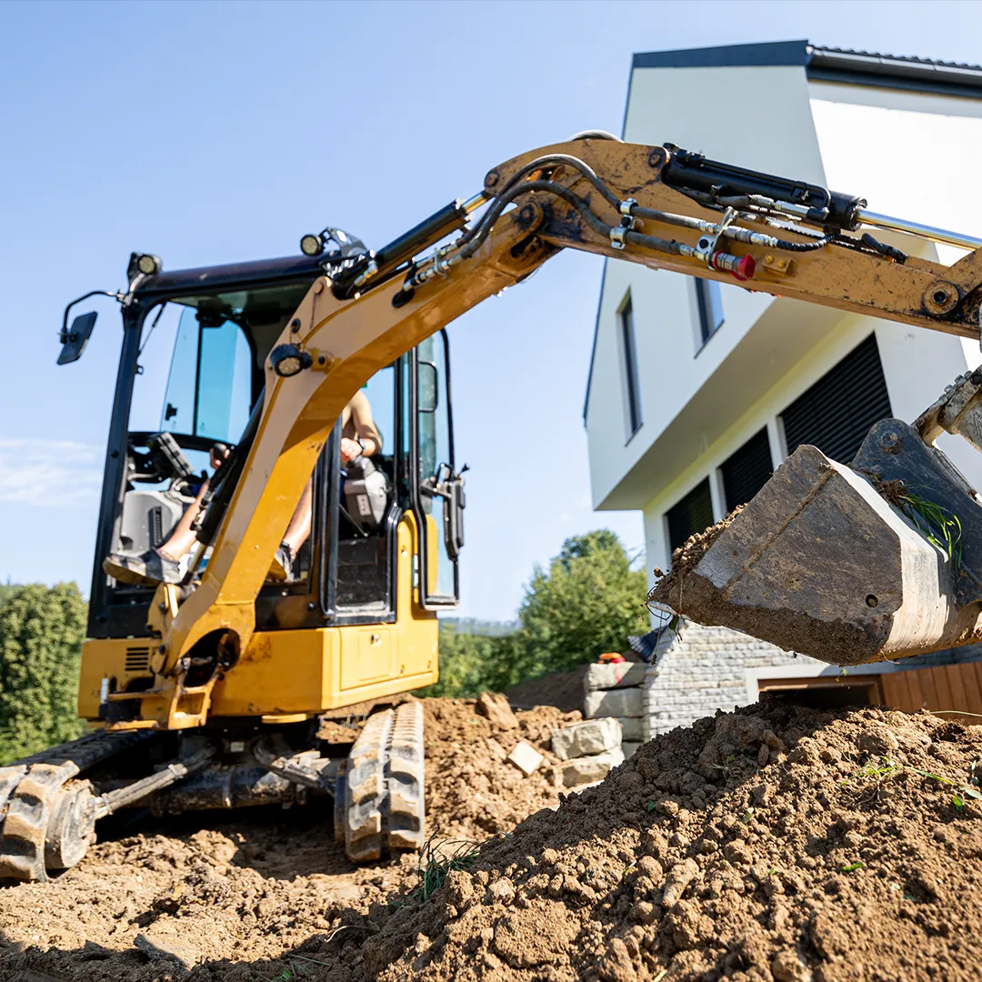 A yellow excavator operates near a modern white house under construction, moving dirt with its bucket as part of the excavation and small demo process. A dumpster removal service is on standby to clear away debris efficiently.