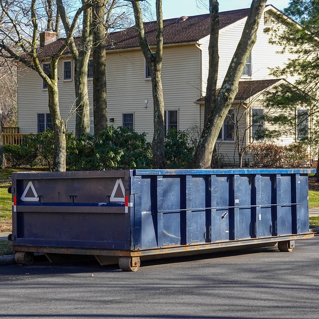 A large blue and black dumpster, likely used for construction or excavation projects, sits on the street in front of a suburban house with trees and shrubs in the background.