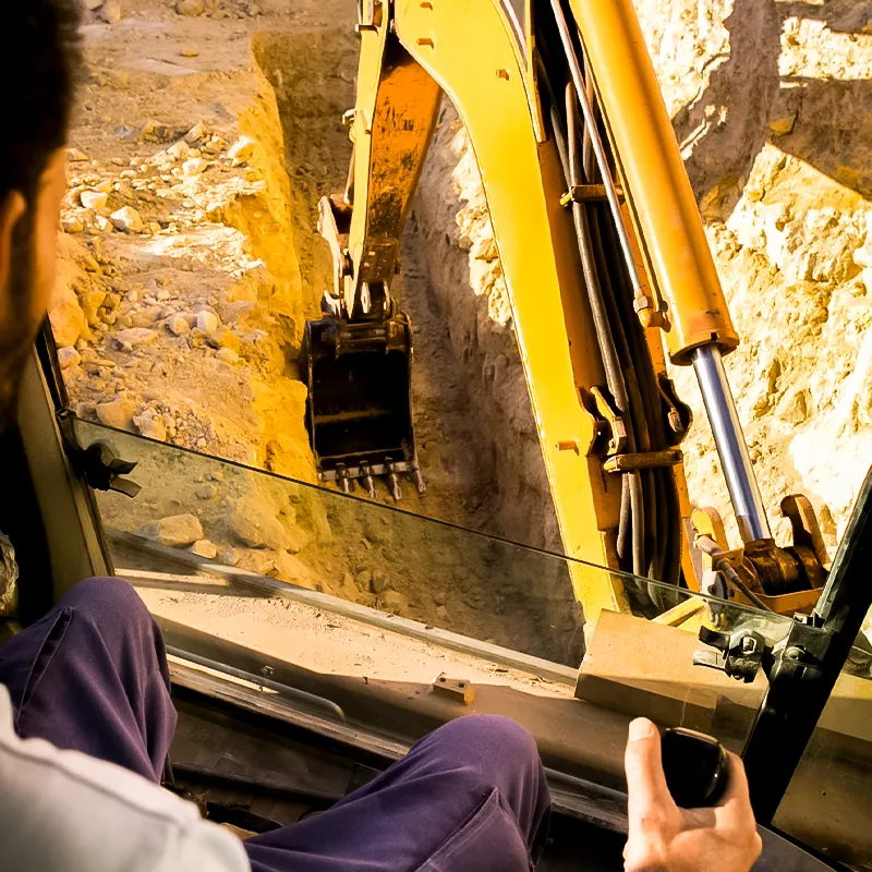 A person operates an excavator, digging a trench in a sandy area, taken from the operator's perspective on the construction site.