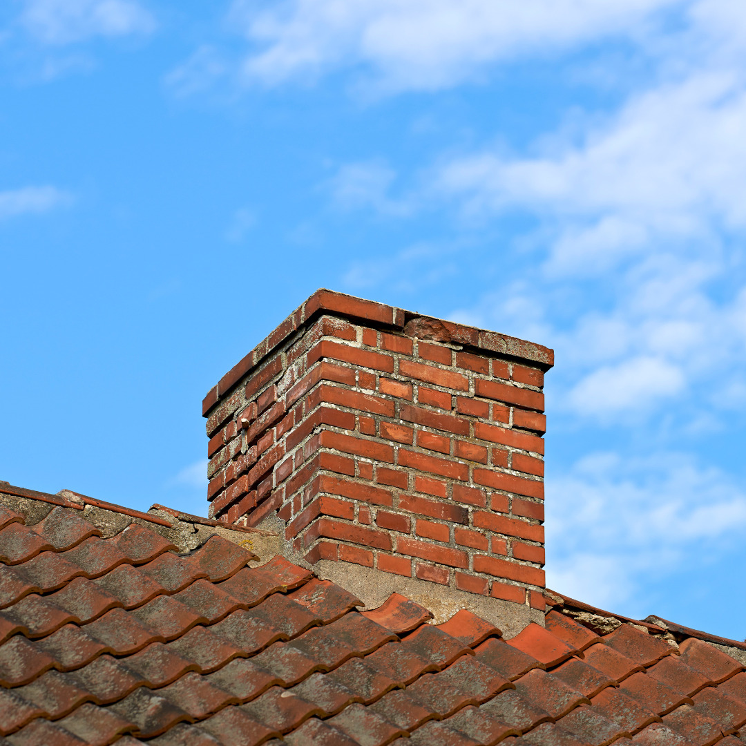 A sturdy brick chimney stands proudly on a slanted tiled roof, set against a blue sky with scattered clouds, awaiting its turn in the rhythm of change that comes with every garage tear down.